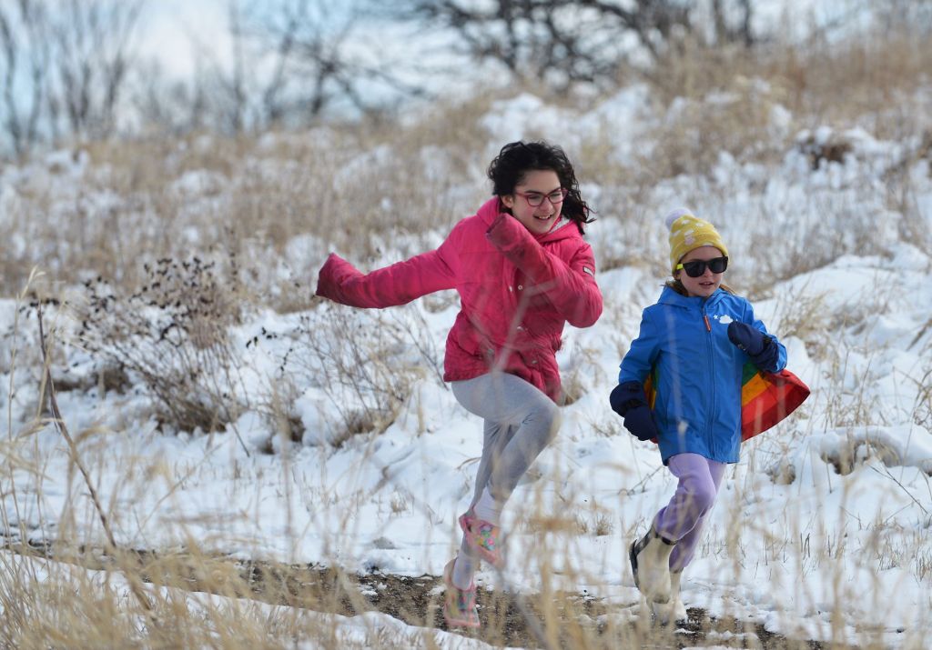 Two young girls in bright pink and blue winter coats joyfully running along a snow-dusted trail at the Prairie Wetlands Learning Center.