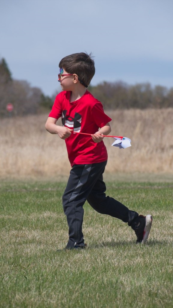 A young boy in a red shirt and black track pants runs across a green field while holding a pinwheel during an outdoor event.
